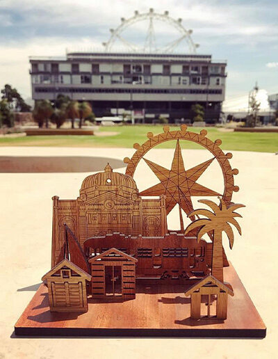 A wooden mini landscape of Melbourne with a tram, bathing houses, a palm tree, Flinders Street Station and the Melbourne Star. Five wooden images sit into a wooden base to make up the 3D landscape.