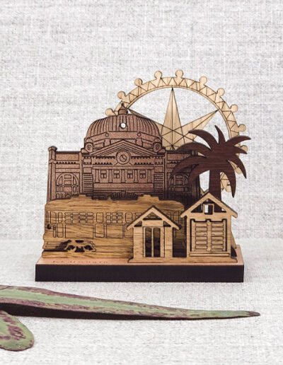 A wooden mini landscape of Melbourne with a tram, bathing houses, a palm tree, Flinders Street Station and the Melbourne Star. Five wooden images sit into a wooden base to make up the 3D landscape.