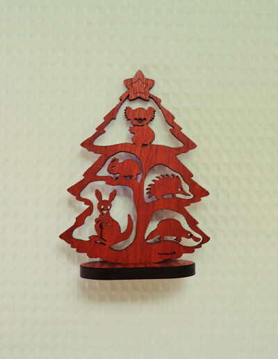 A small wooden Australian made Christmas tree. It is a cut out of a tree with a koala, a wombat, an echidna, a kangaroo and a platypus in it and it stands on a little wooden stand. Its recycled cardboard presentation box is in the foreground